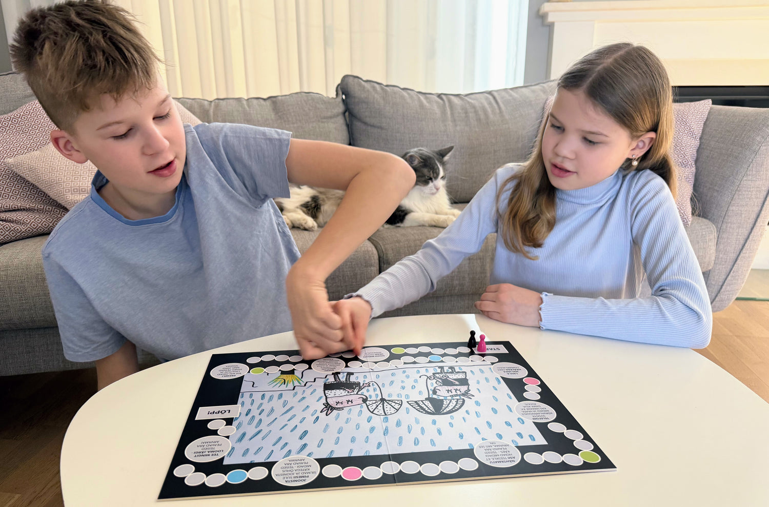 Two children playing a personalized board game on a table, with a cat resting on the couch in the background.