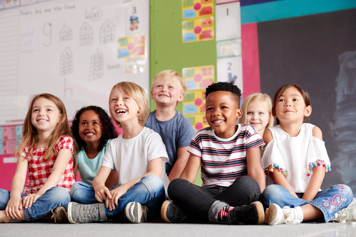 A group of smiling, diverse young children sitting on the floor in a colorful classroom, looking engaged and cheerful