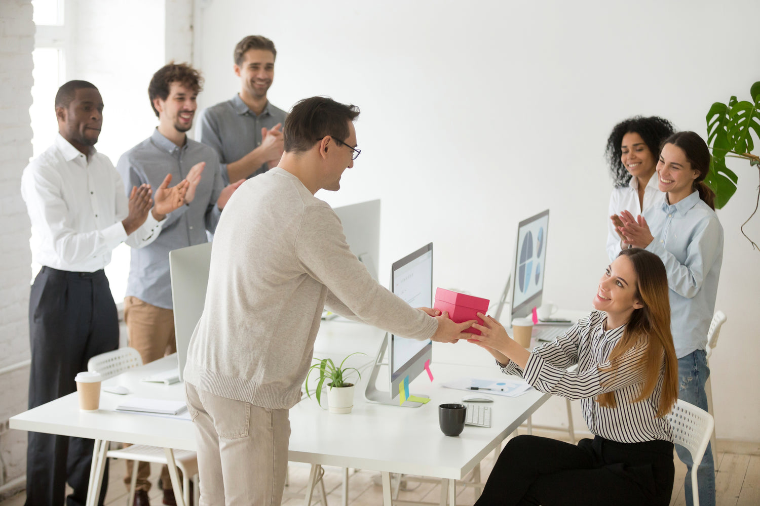 A man giving a gift box to a smiling female colleague at her desk while coworkers applaud in a modern office setting, celebrating employee appreciation or a special occasion.