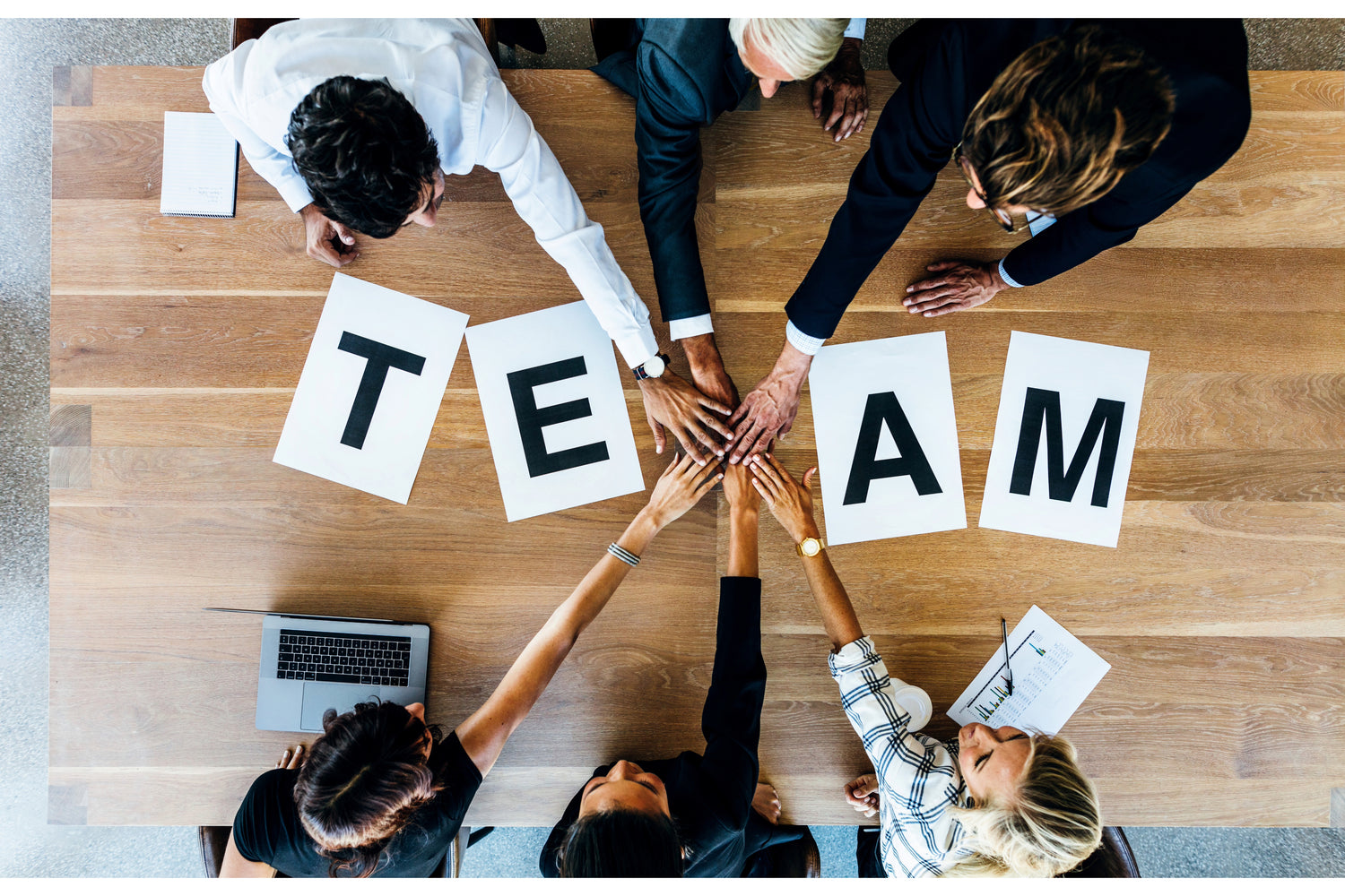 A group of diverse business professionals joining hands over a wooden table with the word "TEAM" spelled out on paper sheets, symbolizing teamwork and collaboration.